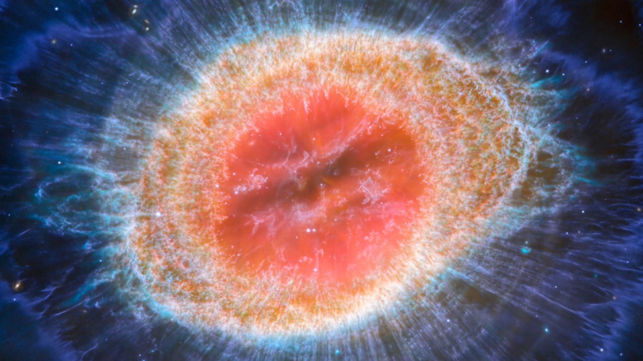 El Telescopio Espacial James Webb de la NASA, la Agencia Espacial Europea y la Agencia Espacial Canadiense ha observado la famosa Nebulosa del Anillo con un detalle sin precedentes.  La Nebulosa del Anillo consiste en una estrella que arroja sus capas exteriores cuando se queda sin combustible y es una nebulosa planetaria típica.  También conocidas como M57 y NGC 6720, ambas están relativamente cerca de la Tierra, a unos 2.500 años luz de distancia.  Esta nueva imagen proporciona una resolución espacial y una sensibilidad espectral sin precedentes.  En particular, el MIRI (instrumento de infrarrojo medio) de Webb revela detalles particulares en las características concéntricas en las regiones exteriores de las nebulosas anulares (derecha).  En la nebulosa hay alrededor de 20.000 esferas densas, que son ricas en hidrógeno molecular.  Por el contrario, en la zona interior aparecen gases muy calientes.  La capa principal contiene un anillo delgado de emisión mejorada de moléculas basadas en carbono conocidas como hidrocarburos aromáticos policíclicos (PAH).  Detrás del borde exterior del anillo principal se encuentran unos diez arcos concéntricos.  Se cree que los arcos surgen de la interacción de la estrella central con una compañera de baja masa que orbita a una distancia similar a la que hay entre la Tierra y el planeta enano Plutón.  De esta manera, nebulosas como la Nebulosa del Anillo revelan una especie de artefacto astronómico, ya que los astrónomos estudian la nebulosa para identificar la estrella que la creó. [Image description: This image of the Ring Nebula appears as a distorted doughnut. The nebula's inner cavity hosts shades of red and orange, while the detailed ring transitions through shades of yellow in the inner regions and blue/purple in the outer region. The ring's inner region has distinct filament elements.]