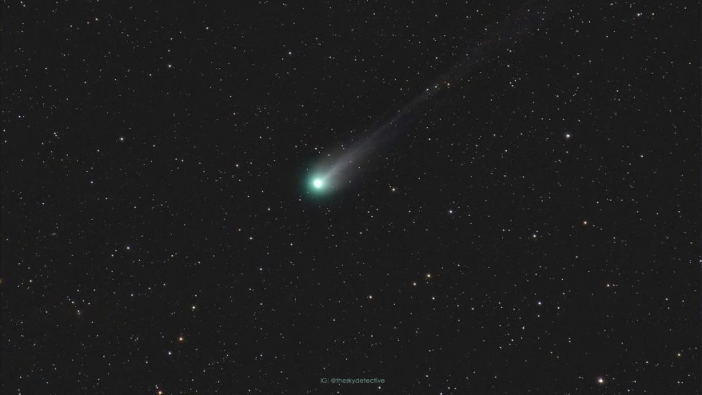 Comet 12P/Pons-Brooks with a glowing green nucleus and long tail streaking behind against a background of stars.